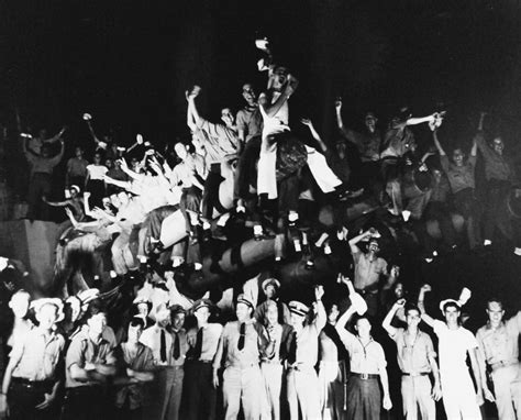 A group of sailors shouting Hoo-Rah, with a naval aircraft carrier in the background